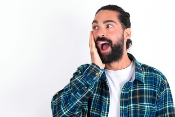 young bearded hispanic man wearing plaid shirt over white background excited looking to the side hand on face. Advertisement and amazement concept.