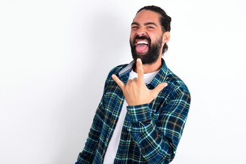 Fototapeta premium Portrait of a crazy young bearded hispanic man wearing plaid shirt over white background showing tongue horns up gesture, expressing excitement of being on concert of band.