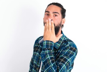 Oh! I think I said it! Close up portrait young bearded hispanic man wearing plaid shirt over white background cover open mouth by hand palm, look at camera with big eyes.