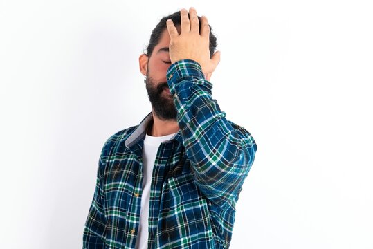 Frustrated young bearded hispanic man wearing plaid shirt over white background holding hand on forehead being depressed regretting what he did having headache, looking stressful.