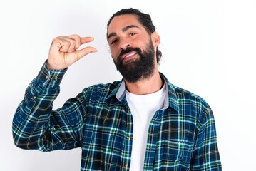 Fototapeta premium Shocked young bearded hispanic man wearing plaid shirt over white background shows something little with hands, demonstrates size, opens mouth from surprise. Measurement concept.