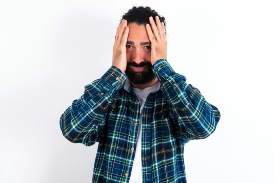 young bearded hispanic man wearing plaid shirt over white background holding head in hands with unhappy expression watching sad movie about animals and trying not to cry.