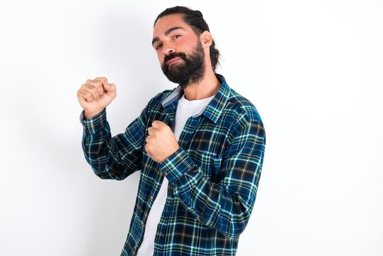 Portrait of attractive young bearded hispanic man wearing plaid shirt over white background holding hands in front of him in boxing position going to fight.