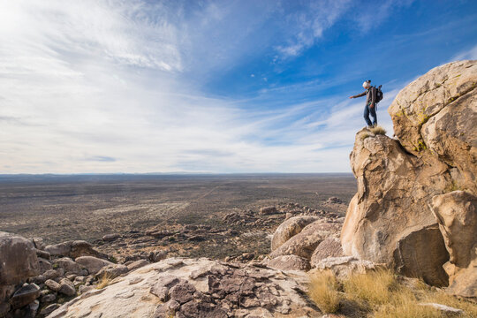 A young, male adventurer, standing on top of a granite cliff, points at something in the distance in the desertic landscape of PeÃ±oles, Chihuahua.