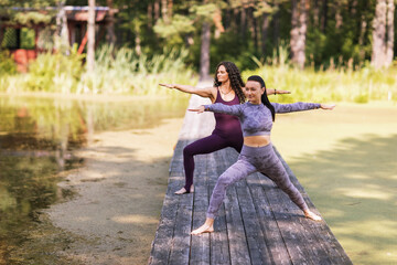 Two women practicing yoga, doing the exercise virabhadrasana, warrior pose, training while standing on a wooden bridge in the park