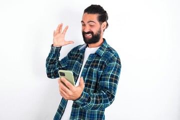 Photo of outraged annoyed young bearded hispanic man wearing plaid shirt over white background holds cell phone, makes call, argues with colleague,  expresses negative emotions. People and anger.