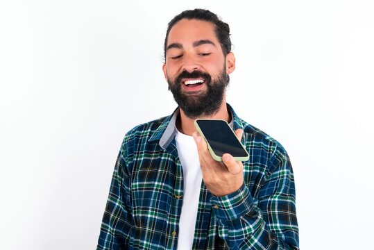 Smiling young bearded hispanic man wearing plaid shirt over white background sending voice message on her smart phone. Communication and new technologies concept.