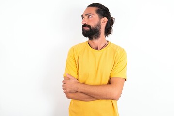 Fototapeta premium Pleased young bearded hispanic man wearing yellow T-shirt over white background keeps hands crossed over chest looks happily aside