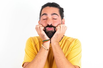 young bearded hispanic man wearing yellow T-shirt over white background with surprised expression keeps hands under chin keeps lips folded makes funny grimace