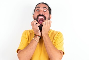 Speechless young bearded hispanic man wearing yellow T-shirt over white background keeps hands near opened mouth reacts to shocking news stares wondered at camera