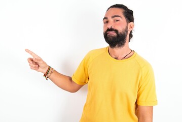 Fototapeta premium young bearded hispanic man wearing yellow T-shirt over white background points at copy space and advertises something, advices best price.