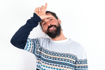 Funny young bearded hispanic man wearing knitted sweater over white background makes loser gesture mocking at someone sticks out tongue making grimace face. - obrazy, fototapety, plakaty