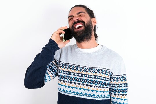 Overemotive happy young bearded hispanic man wearing knitted sweater over white background laughs out positively hears funny story from friend during telephone conversation