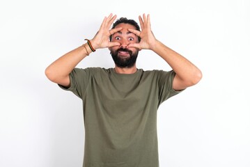 young bearded hispanic man wearing green T-shirt over white background keeping eyes opened to find...