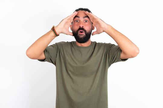 young bearded hispanic man wearing green T-shirt over white background with scared expression, keeps hands on head, jaw dropped, has terrific expression. Omg concept