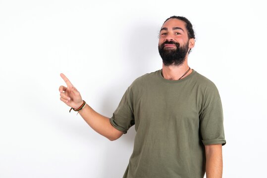Smiling young bearded hispanic man wearing green T-shirt over white background indicating finger empty space showing best low prices