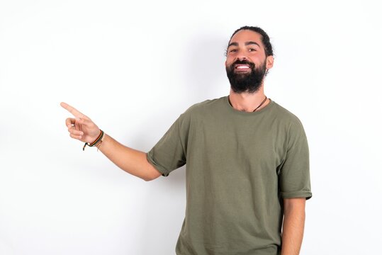 young bearded hispanic man wearing green T-shirt over white background looking at camera indicating finger empty space sales