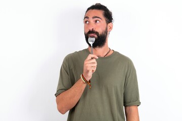 Very hungry young bearded hispanic man wearing green T-shirt over white background holding spoon...