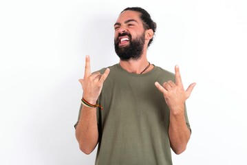young bearded hispanic man wearing green T-shirt over white background making rock hand gesture and...