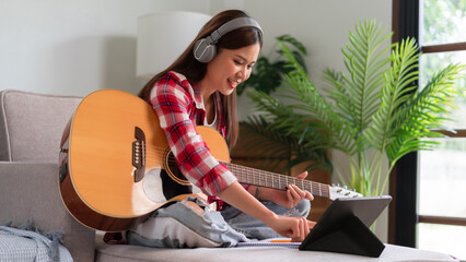 Hobby concept, Young asian woman wear earphone learning music on tablet and playing acoustic guitar