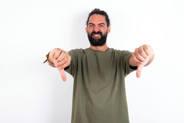 young bearded hispanic man wearing green T-shirt over white background being upset showing thumb down with two hands. Dislike concept.