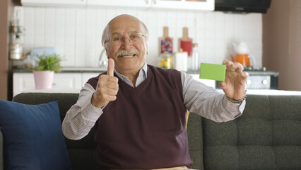 Old man showing green product in hand on sofa, smiling, thumbs up and showing appreciation for...