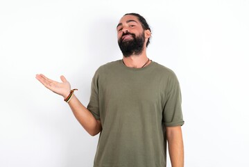 Naklejka premium young bearded hispanic man wearing green T-shirt over white background smiling cheerful presenting and pointing with palm of hand looking at the camera.