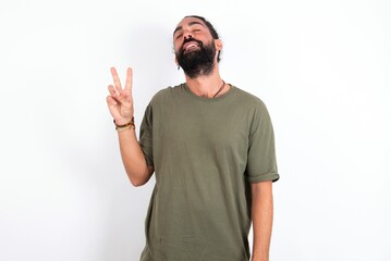 Fototapeta premium young bearded hispanic man wearing green T-shirt over white background smiling with happy face winking at the camera doing victory sign. Number two.