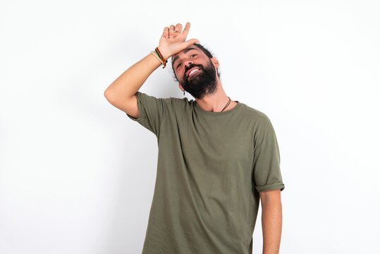 young bearded hispanic man wearing green T-shirt over white background making fun of people with fingers on forehead doing loser gesture mocking and insulting.