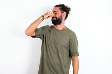 young bearded hispanic man wearing green t-shirt over white background smelling something stinky and disgusting, intolerable smell, holding breath with fingers on nose. Bad smell