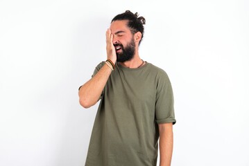 Fototapeta premium young bearded hispanic man wearing green t-shirt over white background Yawning tired covering half face, eye and mouth with hand. Face hurts in pain.