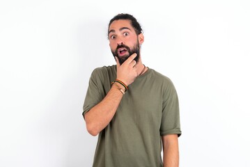 young bearded hispanic man wearing green t-shirt over white background Looking fascinated with disbelief, surprise and amazed expression with hands on chin