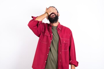 young bearded hispanic man wearing red overshirt over white background smiling and laughing with hand on face covering eyes for surprise. Blind concept.