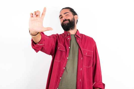 young bearded hispanic man wearing red overshirt over white background making fun of people with fingers on forehead doing loser gesture mocking and insulting.