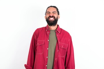 Caucasian man with beard wearing red shirt over white background keeps teeth clenched, frowns face in dissatisfaction, irritated because of much duties.
