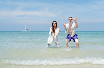 Asian woman with Caucasian man splash water to camera and look fun together with some boat on background on the sea with clear blue sky.