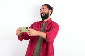 Portrait of an excited Caucasian man with beard wearing red shirt over white background playing games on mobile phone.