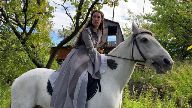 Young woman in image of ancient rider warrior sits on white horse and strokes it in forest.