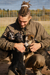 Young Caucasian man with dreadlocks chooses puppy from shelter, sitting in aviary, smiling and rejoicing. Alaskan Husky kennel, volunteer takes care of dogs. Human hugs three puppies.
