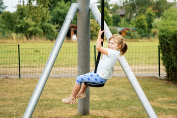 Happy kid girl rids on zip line swing outdoor game play equipment on playground. Child having fun...