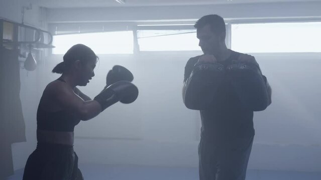 Kickboxing coach and female fighter training leg strike in gym slow motion, teaching attack punch technique wide shot. Fighting instructor and kickboxer silhouettes practicing