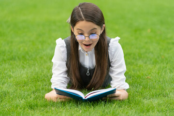 Portrait of surprised girl reading book lying on grass, reading. Teen girl reading outdoors