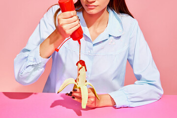 Food pop art photography. Young woman tasting banana with tomato ketchup isolated on pink background. Vintage, retro style interior. Surrealism