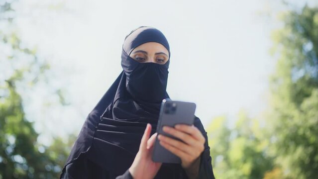 Middle-eastern woman in burqa texting on smartphone outdoor, mobile application
