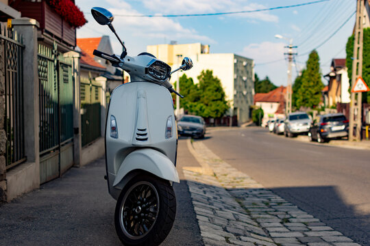 Vintage white motorcycle scooter or moped parked on a sidewalk with empty street, cars and buildings in background