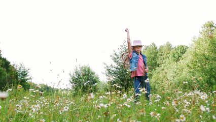 In the grass, among the daisies, in the meadow, dancing, jumping, having fun, a pretty girl, about seven years old. She has long blond hair, is dressed in a denim vest and a pink hat. High quality