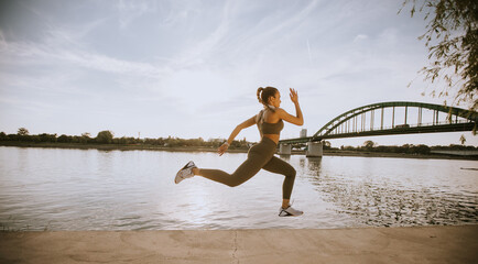 Active young beautiful woman running on the promenade along the riverside - obrazy, fototapety, plakaty