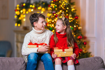 Obraz na płótnie Canvas cheerful children with smile, brother and sister, opening gift boxes tied, sitting on couch at home in evening. children laugh and hold gifts. Christmas tree with light garlands in background