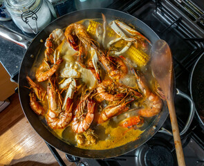 Seafood boil with crabs, prawns and corn on the cob boiling in a wok on a hob in a home kitchen.