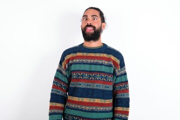 Fototapeta premium Funny Caucasian man with beard wearing sweater over white background makes grimace and crosses eyes plays fool has fun alone sticks out tongue.